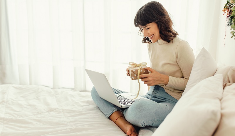 Frau, die auf dem Bett sitzt und einen Videoanruf auf einem Laptop macht, der ihr Ostergeschenk zeigt. Lächelnde Frau, die zu Hause Ostern feiert.