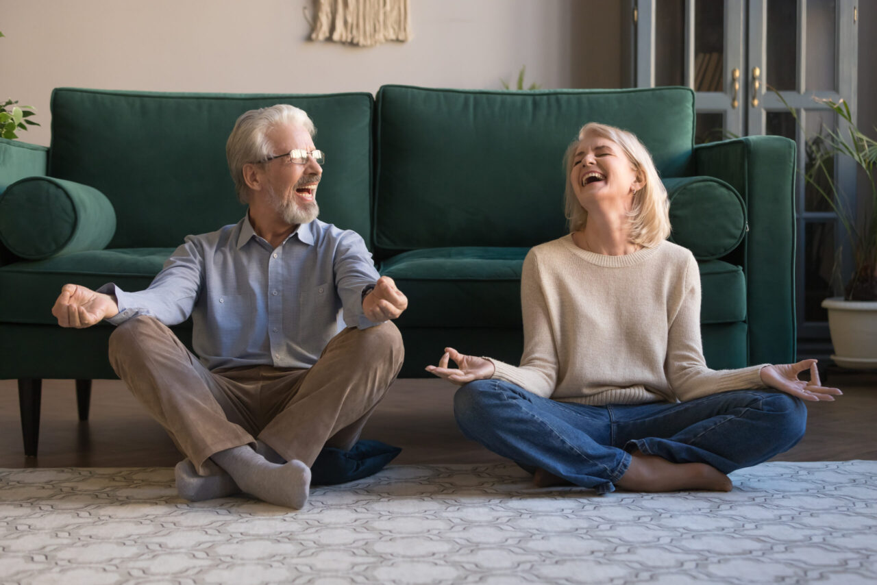 Happy mature couple having fun, practicing yoga together at home, laughing grey haired man and woman sitting in lotus pose on floor in living room, breathing, relaxing, healthy lifestyle concept